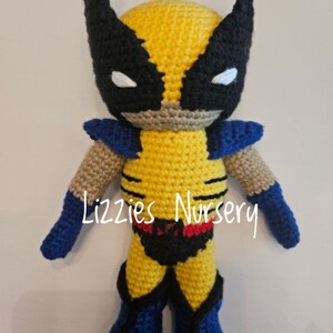 May include: A crocheted stuffed toy of Wolverine from the X-Men. The toy is yellow with black claws, a black mask with white eyes, and blue boots. The toy is standing on a white surface. The text "Lizzie's Nursery" is visible on the toy's chest.