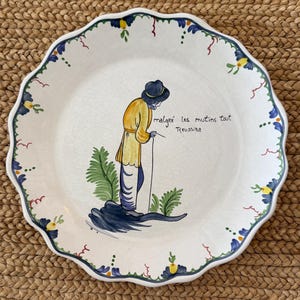 May include: A white ceramic plate with a scalloped edge, featuring a hand-painted illustration of a person in a yellow coat and black hat. The plate has a blue and green floral border and the text "malgré les mutins tout Reussira".