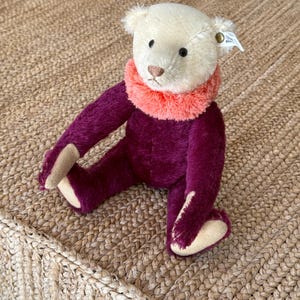 May include: A small, cream-colored teddy bear wearing a purple outfit with a pink fur collar. The bear is sitting on a woven rug.