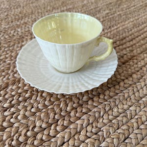 Puede incluir: Un juego de taza y platillo blancos. La taza tiene un exterior texturizado y un interior y asa de color amarillo pálido. El platillo tiene un borde festoneado. El juego está sobre una superficie tejida de color canela.