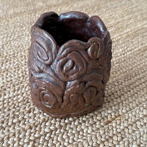 Puede incluir: Un jarrón de cerámica marrón hecho a mano con una superficie texturizada. El jarrón presenta un diseño floral repetido alrededor del exterior. El interior es oscuro y el jarrón se encuentra sobre una superficie tejida de color beige. El jarrón mide unos 10 cm de alto.