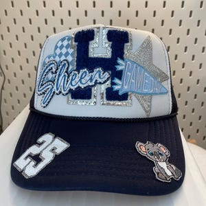 May include: A navy blue and white trucker hat with the word "Sheen" in blue script. The hat features a large blue "H" and a silver star. A koala patch and the number "23" are on the brim. The words "Game Day" are also on the hat.