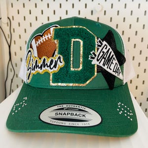 May include: Green and white snapback hat with a football and the word "Zimmer" in gold and white script. The letter "D" is in the center, with a "Game Day" patch. The hat has a white mesh back and a curved brim.
