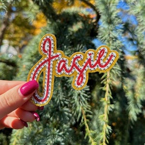 May include: A glittery, cursive "Jazzie" patch with a gold border, red lettering, and silver rhinestone accents. The patch is held against a backdrop of green foliage.
