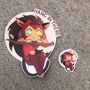 K&ouml;nnte beinhalten: Zwei Aufkleber mit einer Comic-Katzenfigur mit roten Haaren, gelben Augen und einer rosa Nase. Der gr&ouml;&szlig;ere Aufkleber ist ein runder Aufkleber mit dem Text "Hang in there!" Der kleinere Aufkleber ist ein Kreis mit dem gleichen Text.