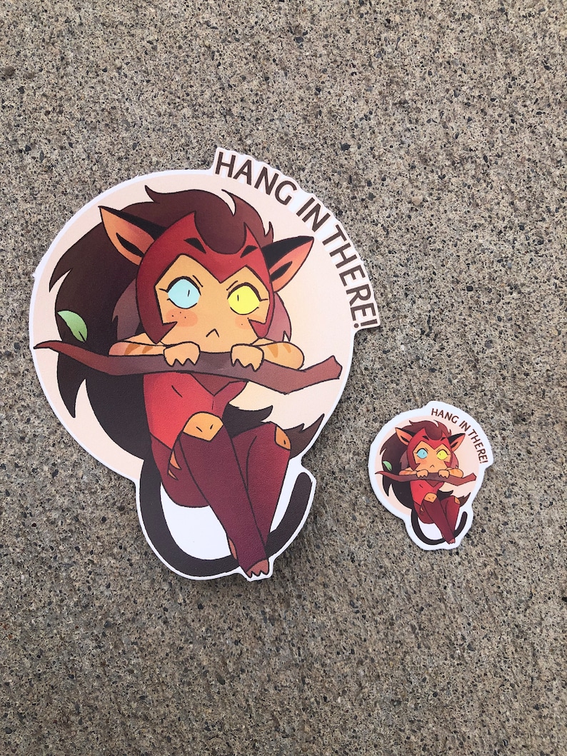 K&ouml;nnte beinhalten: Zwei Aufkleber mit einer Comic-Katzenfigur mit roten Haaren, gelben Augen und einer rosa Nase. Der gr&ouml;&szlig;ere Aufkleber ist ein runder Aufkleber mit dem Text "Hang in there!" Der kleinere Aufkleber ist ein Kreis mit dem gleichen Text.