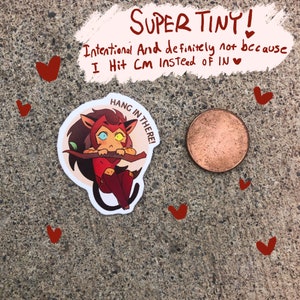 K&ouml;nnte beinhalten: Ein kleiner, runder Aufkleber mit einer Comic-Katze mit rot-orangem Fell, gelben Augen und einem wei&szlig;en Stirnband. Die Katze h&auml;lt sich an einem Ast fest und der Aufkleber sagt "Hang in there!" in einer wei&szlig;en Sprechblase.