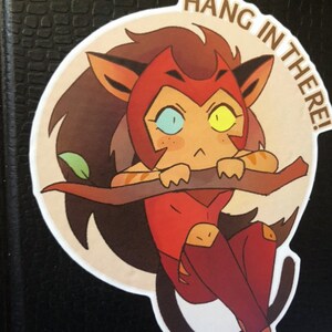 K&ouml;nnte beinhalten: Ein runder Aufkleber mit einem Cartoon-Bild eines rot-braunen Katers mit gelben und blauen Augen. Die Katze h&auml;lt sich mit ihren Pfoten an einem Ast fest. Der Text "HANG IN THERE!" ist in einem Kreis um die Katze geschrieben.