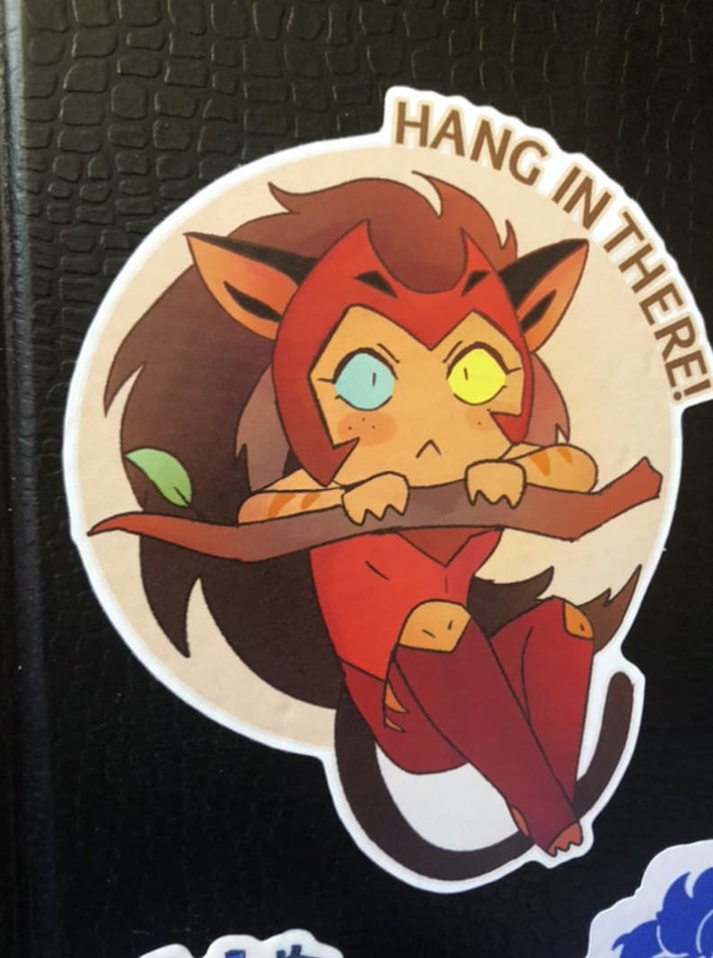 K&ouml;nnte beinhalten: Ein runder Aufkleber mit einem Cartoon-Bild eines rot-braunen Katers mit gelben und blauen Augen. Die Katze h&auml;lt sich mit ihren Pfoten an einem Ast fest. Der Text "HANG IN THERE!" ist in einem Kreis um die Katze geschrieben.
