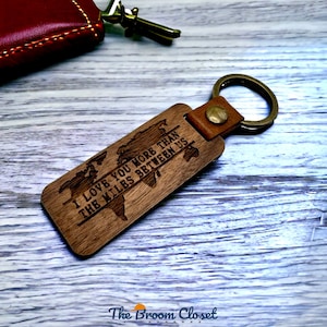 May include: A rectangular wooden keychain with a world map design and the engraved text "I LOVE YOU MORE THAN THE MILES BETWEEN US." The keychain has a leather strap and a metal ring. The Broom Closet logo is at the bottom.