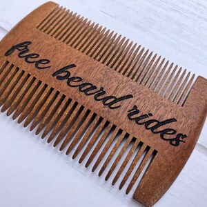 May include: A wooden beard comb with the words "free beard rides" etched in black script. The comb has two sides with fine teeth for grooming. The wood has a warm brown tone.