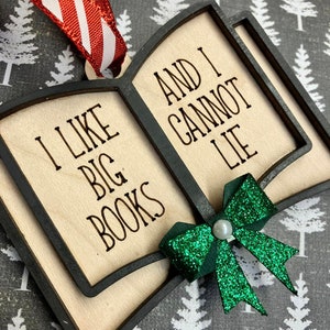 May include: Wooden book-shaped ornament with the text "I LIKE BIG BOOKS AND I CANNOT LIE". The ornament has a black frame, a green glitter bow, and a red and white striped ribbon for hanging. The background is a gray and white tree pattern.