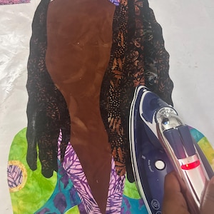 May include: A person is ironing a fabric piece with a blue and silver iron. The fabric is a colorful patterned design with a brown fabric piece on top. The iron has a "Auto Clean" button and a light on the side.