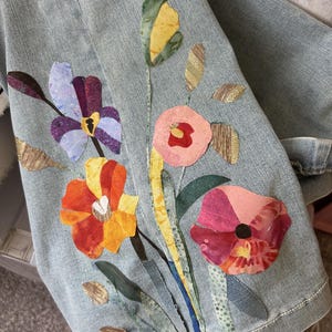 Könnte beinhalten: Hellblaue Jeansjacke mit farbenfrohen Blumenapplikationen. Die Blumen haben orange, gelbe, violette und rosa Blütenblätter mit grünen und goldenen Akzenten. Der Ärmel der Jacke ist sichtbar und zeigt die detaillierte Verarbeitung.