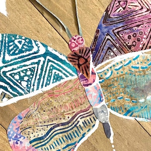 May include: A colorful butterfly with wings made of patterned fabric. The butterfly is a collage of different fabrics with geometric and floral designs. The wings are a mix of teal, purple, pink, and brown.
