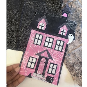 Peut inclure: Décoration de maison rose et noire sur le thème d'Halloween avec un design effrayant. La maison présente des toiles d'araignées blanches, des fenêtres et un fantôme. Un chapeau de sorcière et un chaudron sont également visibles. La maison est tenue par une main.