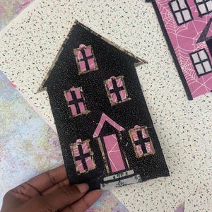 May include: Black fabric house-shaped applique with pink window and door accents. The house has a glittery texture and is set against a speckled background. Other similar house appliques are visible.