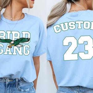 A woman wearing a light blue t-shirt with the text "BIRD GANG" and the number "23" printed on it.