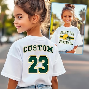 May include: White t-shirt with "CUSTOM" and the number "23" in green and gold lettering on the back. The front of the shirt in the image has "GREEN BAY FOOTBALL" with a helmet graphic.