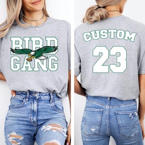 A woman wearing a grey t-shirt with the text "BIRD GANG" and the number "23" printed on it, along with a graphic of an eagle holding a football.