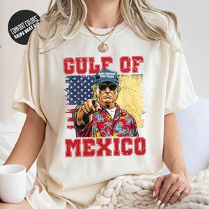 A person wearing a white t-shirt with a graphic design depicting a man in a Hawaiian shirt and sunglasses, pointing towards the text "GULF OF MEXICO" and the American flag.