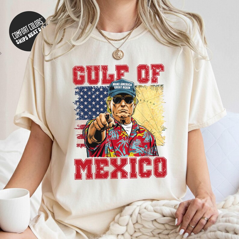 A person wearing a white t-shirt with a graphic design depicting a man in a Hawaiian shirt and sunglasses, pointing towards the text "GULF OF MEXICO" and the American flag.