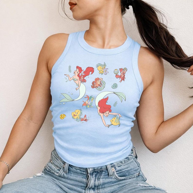 A woman wearing a light blue tank top with a mermaid design and a ponytail.