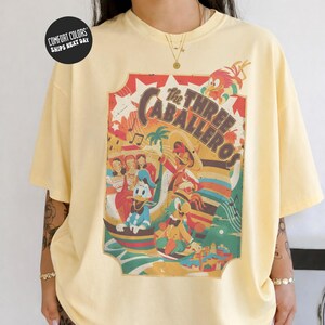 A person wearing a yellow t-shirt with a colorful graphic design featuring cartoon characters and text.
