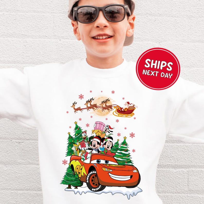 A young boy wearing a white sweater with a Christmas-themed design featuring characters from the Disney movie Cars and a Santa Claus sleigh.