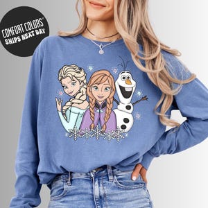 A woman wearing a blue long-sleeved shirt with a graphic of three cartoon characters - two girls and a snowman - on it.