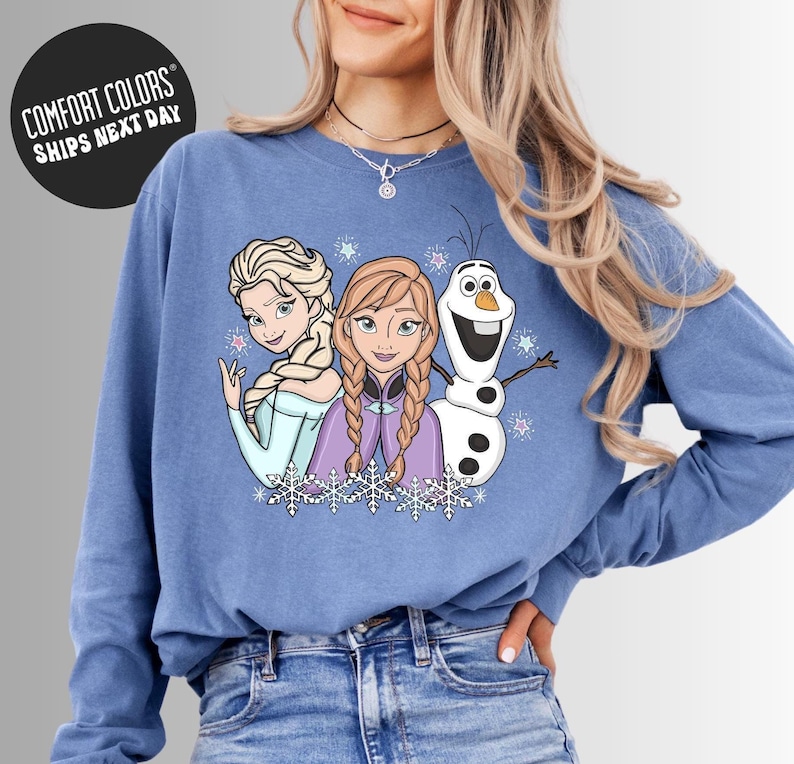 A woman wearing a blue long-sleeved shirt with a graphic of three cartoon characters - two girls and a snowman - on it.