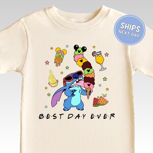 A white t-shirt with a colorful graphic design featuring a cartoon character holding a variety of ice cream cones and other desserts, surrounded by stars and other whimsical elements. The text "BEST DAY EVER" is displayed below the character.