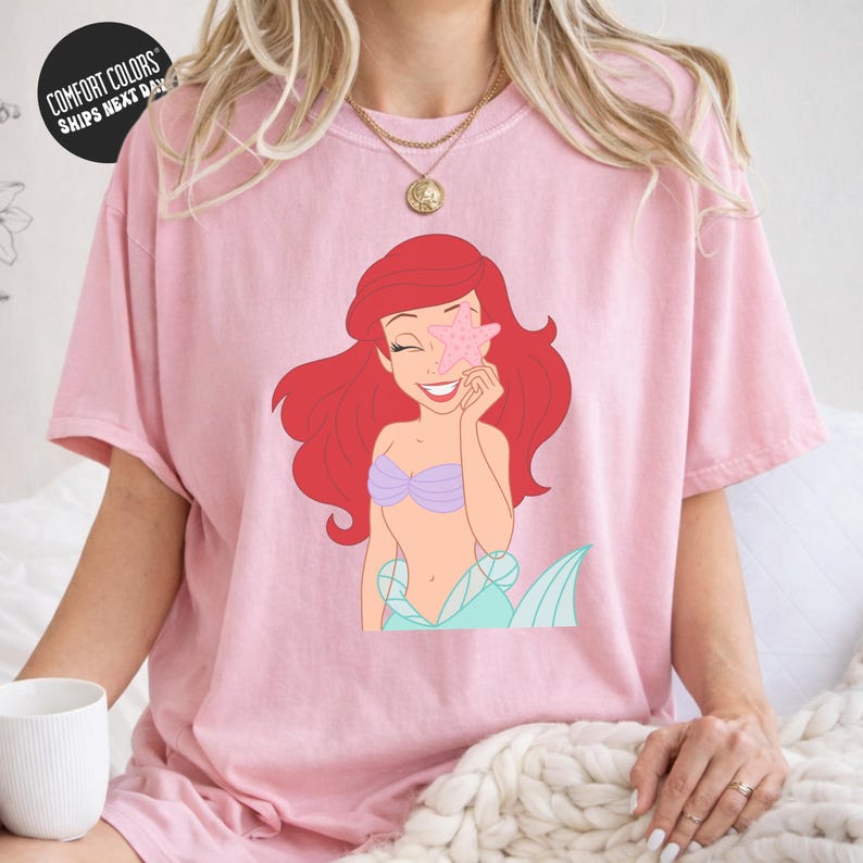 A woman wearing a pink t-shirt with a cartoon image of a mermaid character.