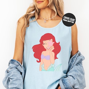 A woman wearing a blue tank top with a cartoon image of a mermaid and a necklace.