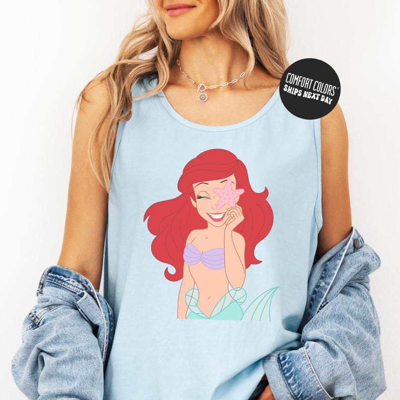 A woman wearing a blue tank top with a cartoon image of a mermaid and a necklace.