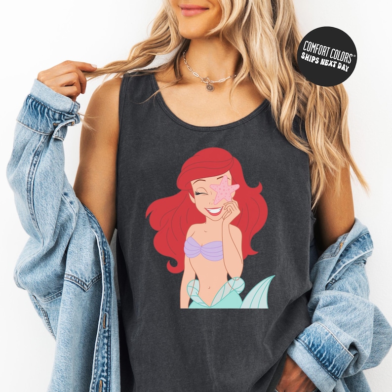 A woman wearing a black tank top with a graphic of a mermaid character from the Disney film "The Little Mermaid". She is also wearing a denim jacket and has her hair styled in loose curls.
