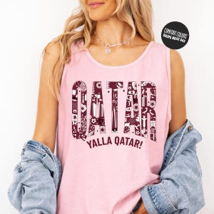 A woman wearing a pink tank top with the word "QATAR" printed on it, along with the phrase "YALLA QATAR!" printed below. She is also wearing a denim jacket and has her arms crossed.