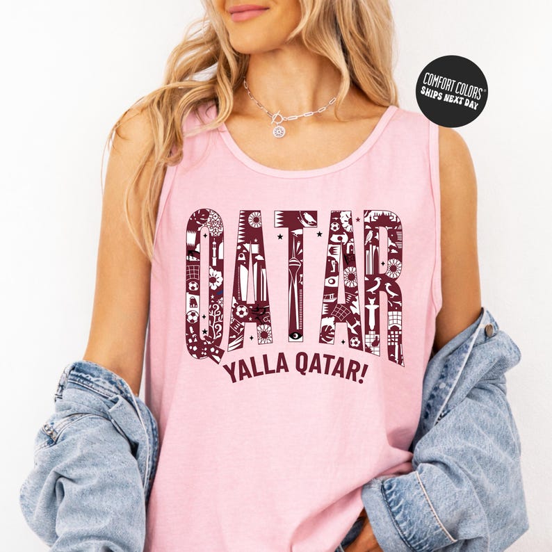 A woman wearing a pink tank top with the word "QATAR" printed on it, along with the phrase "YALLA QATAR!" printed below. She is also wearing a denim jacket and has her arms crossed.