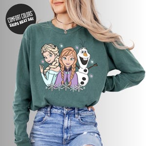 A woman wearing a green long-sleeved shirt with a graphic of three cartoon characters - two women and a snowman - on it.