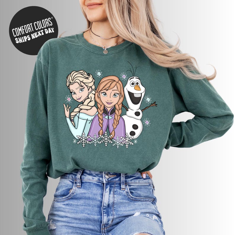 A woman wearing a green long-sleeved shirt with a graphic of three cartoon characters - two women and a snowman - on it.