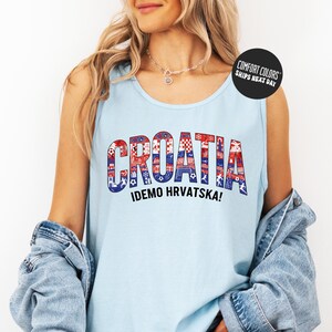 A woman wearing a light blue tank top with the word "Croatia" printed on it, along with the phrase "IDEMO HVRVATSKA!" printed below. She is also wearing a denim jacket and has a necklace around her neck.