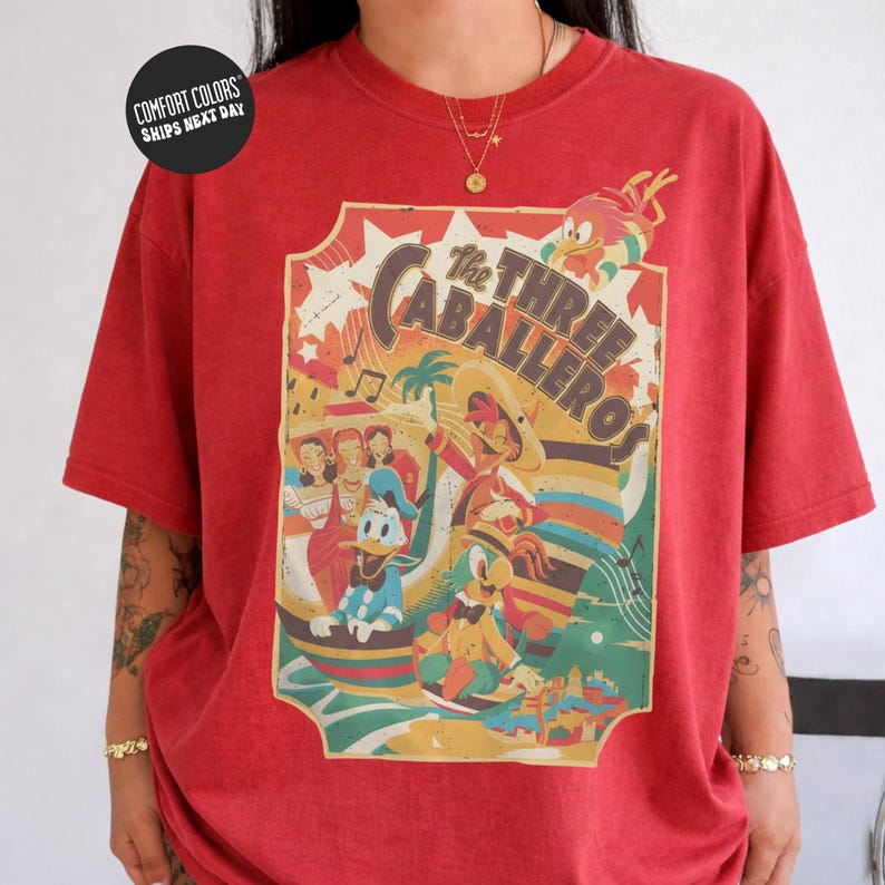 A red t-shirt with a colorful graphic design featuring cartoon characters and various elements, including a large sunburst and a palm tree.