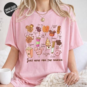 A woman wearing a pink t-shirt with a graphic design of various Disney-themed snacks and treats, including popcorn, cotton candy, ice cream, and cookies.