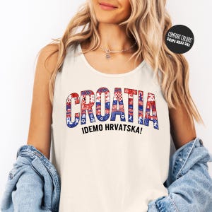 A woman wearing a white tank top with the word "Croatia" printed on it, along with the text "IDEMO HRVATSKA!" The woman is also wearing a denim jacket and has her hands in her pockets.