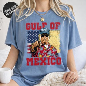 A person wearing a blue t-shirt with a graphic design depicting a man in a Hawaiian shirt and sunglasses, pointing at the viewer, with the text "GULF OF MEXICO" and "MAKE AMERICA GREAT AGAIN" printed on the shirt.