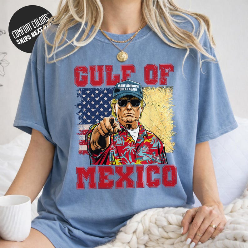 A person wearing a blue t-shirt with a graphic design depicting a man in a Hawaiian shirt and sunglasses, pointing at the viewer, with the text "GULF OF MEXICO" and "MAKE AMERICA GREAT AGAIN" printed on the shirt.