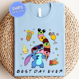 A light blue t-shirt with a colorful graphic design featuring a cartoon character holding a stack of ice cream cones, surrounded by various desserts and stars.