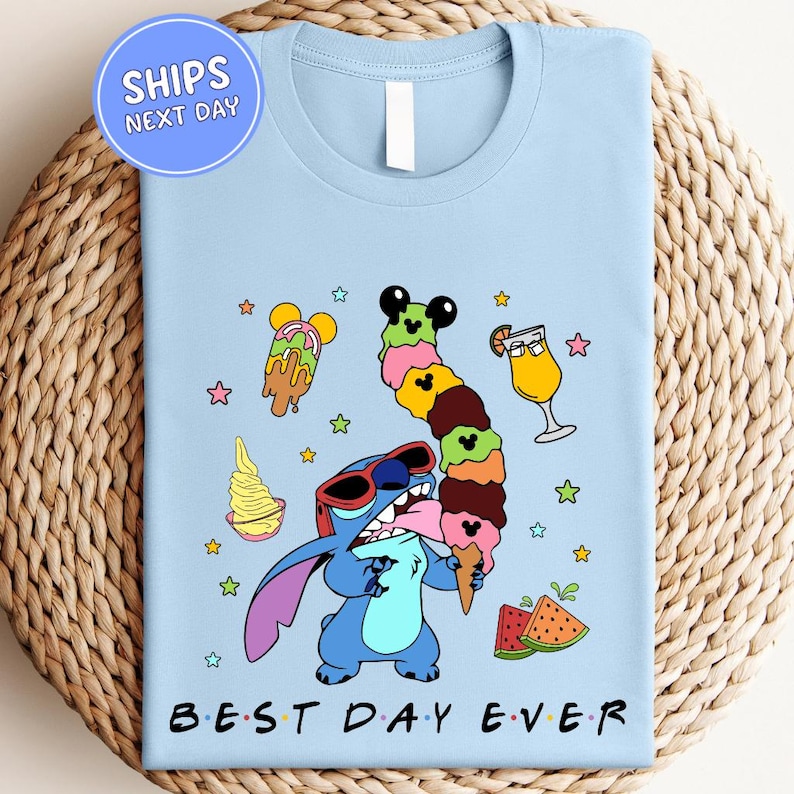 A light blue t-shirt with a colorful graphic design featuring a cartoon character holding a stack of ice cream cones, surrounded by various desserts and stars.