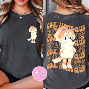 May include: Dark gray t-shirt featuring a cartoon dog with a ponytail. The front of the shirt has a small graphic of the dog, while the back displays the text "COOL AUNTS CLUB" in orange. The shirt is made of a soft material.