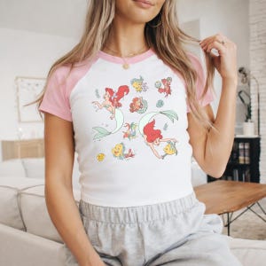A woman wearing a white and pink t-shirt with a mermaid design and a gold necklace.